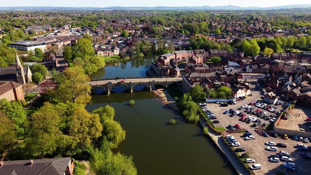 Static aerial drone shot of Welsh Bridge over River Severn, Shrewsbury. Cars crossing stone arch bridge beside church, riverside car park and red brick town panorama.