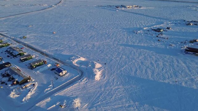 Drone follows parent and child on skidoos (snowmobiles) outside Arviat, Nunavut. Long shadows at golden hour highlight winter travel and Inuit family life on the Arctic tundra.