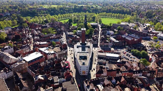 Drone pulling back from Shrewsbury town centre clock tower revealing medieval street layout, red brick rooftops, market square and Tudor buildings on sunny spring day.
