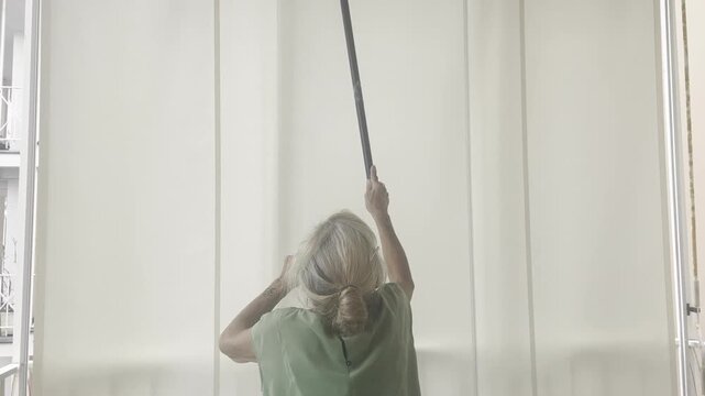 View through a large glass window of an elderly woman in a green shirt, diligently using a pole squeegee to clean high glass. The scene has a soft, reflective, and residential light