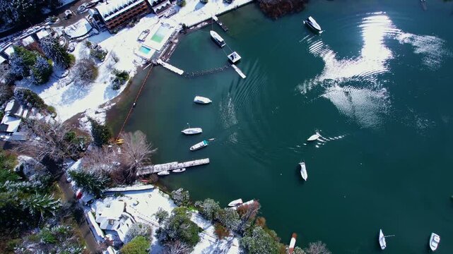 Static drone shot of a boat arriving at a wooden pier, creating ripples in the transparent turquoise water of Nahuel Huapi Lake. Snow-covered shore and sun reflections in winter.