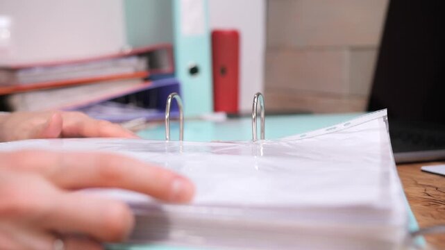 Person's hands organizing important documents within a clear plastic sleeve in a ring binder on a wooden desk, symbolizing paperwork, record keeping, and office administration tasks