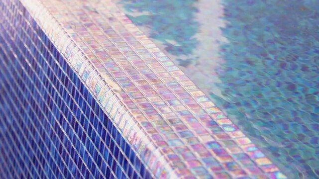 Close-up of water overflowing the edge of an infinity pool with iridescent blue mosaic tiles. Shimmering water surface and luxury pool detail in a high-end resort during a sunny day.
