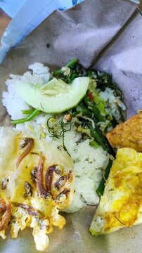 Authentic Indonesian Nasi Pecel with Omelette and Anchovy Peanut Cracker Served on Traditional Brown Paper Wrapper, Healthy Javanese Vegetable Salad Rice Concept for Street Food Industry