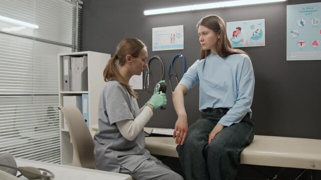 Young doctor taking dermatoscope off medical table and examining arm skin of gen Z patient during dermatological examination