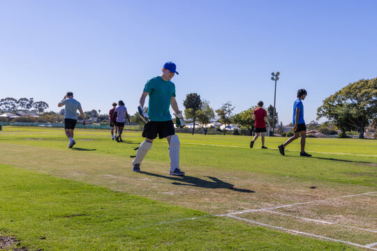 Male teammates walking across marked cricket pitch in park wearing batting pads and gloves