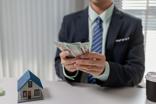 Businessman counting cash with property model and financial report on desk, representing real estate income, investment return, budgeting, and financial management in property business.