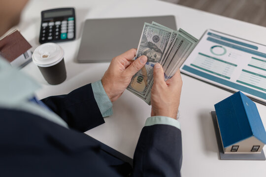 Businessman counting cash with property model and financial report on desk, representing real estate income, investment return, budgeting, and financial management in property business.