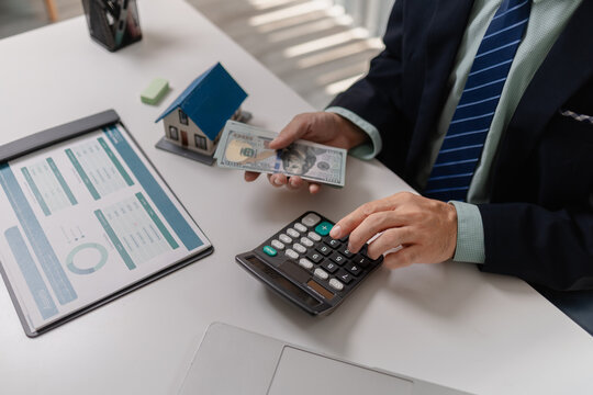 Businessman counting cash with property model and financial report on desk, representing real estate income, investment return, budgeting, and financial management in property business.