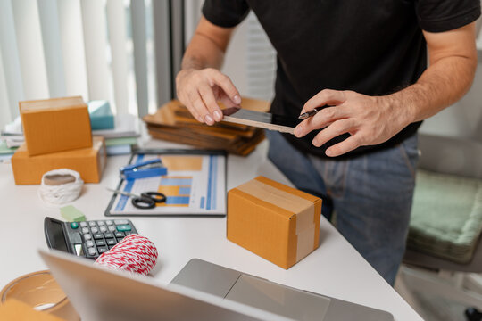 Small business owner using smartphone to check order details before shipping parcel, managing online sales and delivery process, representing ecommerce operations and modern retail workflow.