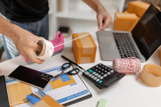 Small business owner tying parcel with string while preparing package for delivery, organizing shipping process and handling ecommerce orders, representing logistics and packaging workflow.