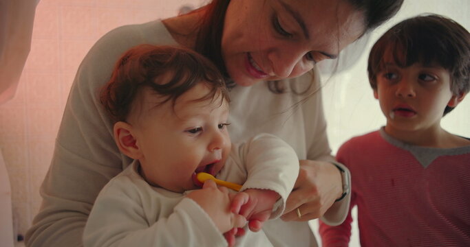 Mother with baby and son in bathroom, smiling gently while helping infant explore toothbrush, warm family interaction showing affection, patience, and nurturing care