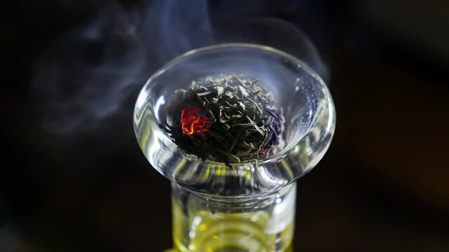 Ecu macro video of hand igniting herbs in glass bong with orange lighter. Red ember, grey smoke to bokeh. Chiaroscuro. Intimate ritual