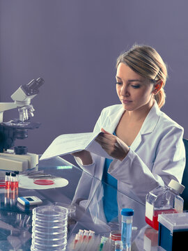 Young european female scientist reading lab notebook beside compound microscope and T-flask