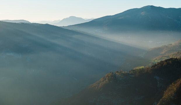 Mountain landscape with sun rays piercing through morning mist over forested valleys and distant peaks, offering a serene tourism destination in Luserna, Alpe Cimbra, Trentino, Italy.