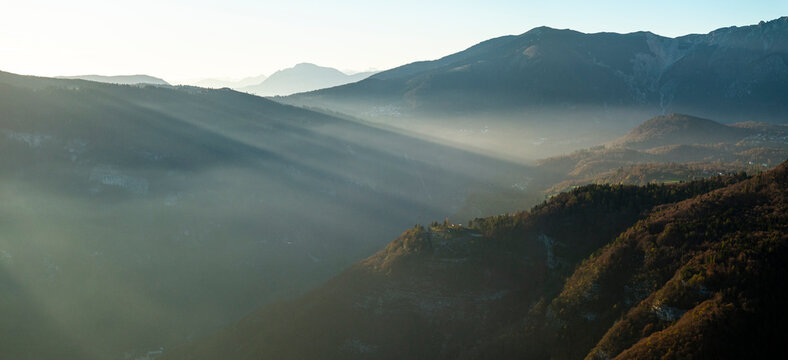 Alpine mountains and forested valleys shrouded in morning mist with sun rays piercing through, creating a serene natural landscape for tourism, Luserna, Alpe Cimbra, Trentino, Italy.