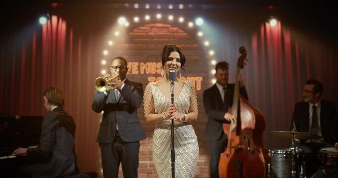 Elegant Female Singer in a Sparkling Gown Performs With a Live Jazz Band in a Cozy Club, as Trumpet, Piano, Bass, Drums and Saxophone Energize the Stage Before Audience. Classical Retro Concert
