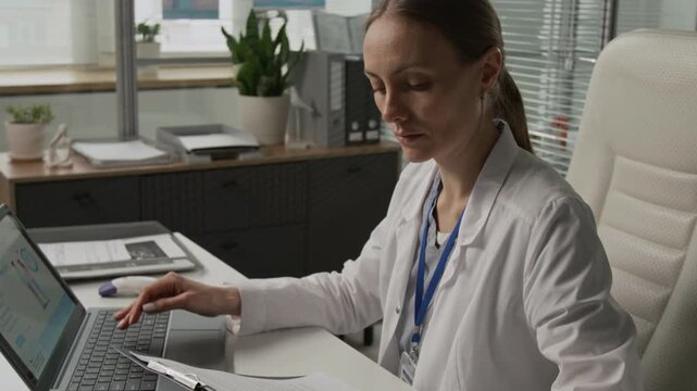 Tilt up view of fertility specialist dressed in lab coat studying medical contract and typing on laptop while sitting at white desk in office chair