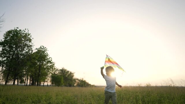 Young boy running across vast summer field. Child enjoys flying bright colorful kite. Golden sunset glows. Happy boy loves running outdoors. Rainbow kite trails above. Nature surrounds calm child.