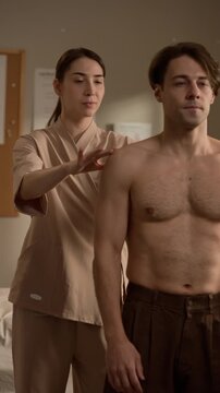 Vertical medium shot of young Caucasian female therapist evaluating shoulder alignment of shirtless male patient raising his arm in wellness room during orthopedic consultation