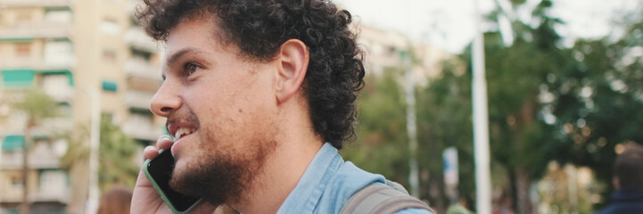 Fototapeta premium Smiling guy dressed in denim shirt with paper bag with healthy food in his hands crosses pedestrian crossing, talking on smartphone. Panoramic