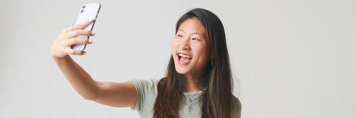 Fototapeta premium Smiling girl in gray top takes selfie on mobile phone, makes video call, isolated on white background, studio shot. Panoramic