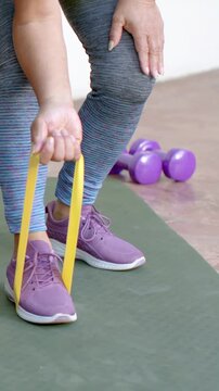 Vertical video: Placing yellow band under right foot, senior in leggings pulling on mat for rehab