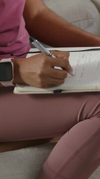 Vertical video: Pen touching lined page, woman in pink writing notes on sofa, shot revealing watch