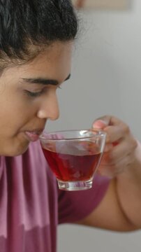 Vertical video: Picking up glass cup, woman in mauve top tasting red herbal tea at home, copy space