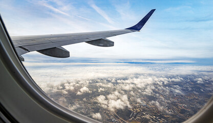 Obraz premium A view from an airplane window shows a silver wing soaring over a landscape of fluffy white clouds, dark blue water, and a coastal town.