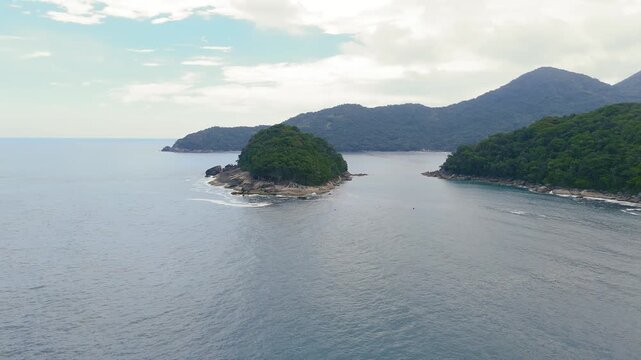 Aerial view of small forested rocky island with mountains and calm ocean water in Trindade, Paraty, Brazil. Orbit camera movement