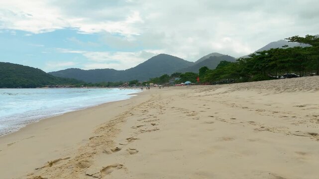 Ground level view of Trindade Beach with footprints in sand, waves and mountains in Paraty, Brazil. Fast push in at low altitude with rain drops on lens