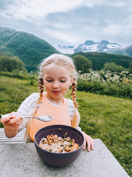 Child girl eating granola breakfast summer picnic outdoor travel camping vacations in Norway, healthy lifestyle vegan healthy food 5 years old kid with pigtails enjoying cereal bowl with a view