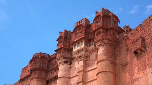 A low-angle static shot of the impressive red sandstone palace structures and towering bastions of Mehrangarh Fort against a bright blue sky in Jodhpur, Rajasthan, India.