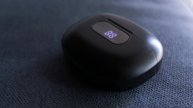 Small black wireless earbuds charging case placed on soft blue fabric with natural daylight and shallow depth of field. Minimal technology and portable audio concept.