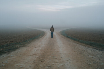 Naklejka premium Misty morning blankets a barren, split dirt path. Lone figure walks forward, unaware of the choice ahead. Dusk's gray light softens the horizon's edge