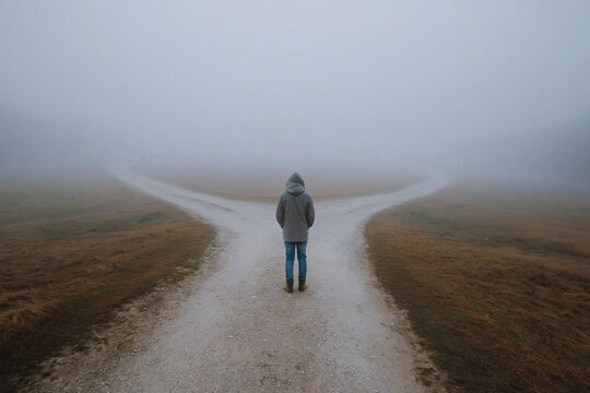 Fog shrouds a split path ahead. One person stands, facing choice. Damp earth and mist fill the air. Gray coat and hood frame the figure. Decision looms in silent uncertainty