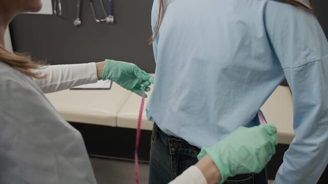 Tilt up shot of positive young doctor measuring waist of long-haired gen Z girl and speaking with her while running appointment in clinic