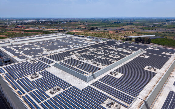 Aerial view of massive solar panel array covering entire rooftop of large industrial warehouse complex surrounded by agricultural fields and rural landscape