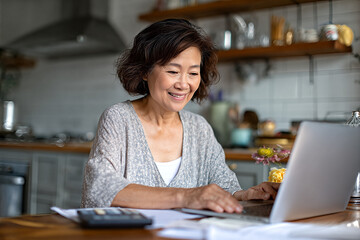 Fototapeta premium Mature Asian remote worker smiling in her kitchen, typing on a laptop amid bills, calculator and a vase of flowers on a wooden table