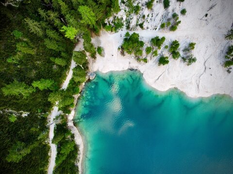 Aerial view of Lake Braies in the Dolomites, Italy, showcasing its turquoise waters adjacent to a light-colored, rocky shore. The surrounding dense forest, primarily composed of conifers like pines 