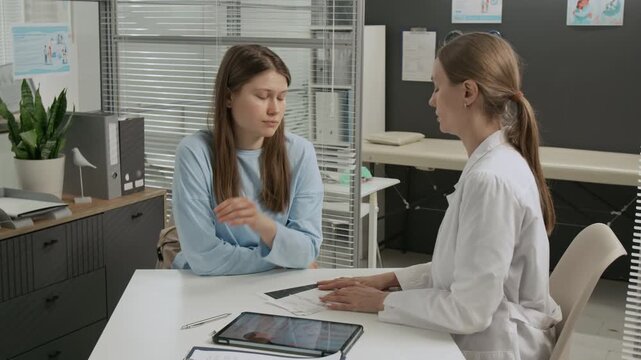 Female gynecologist discussing test results with upset young woman and comforting her by putting hand on patients shoulder during consultation
