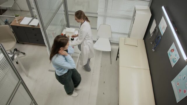 View from above of female doctor and patient entering examination room, girl sitting down on couch, woman putting on gloves and taking stethoscope off wall