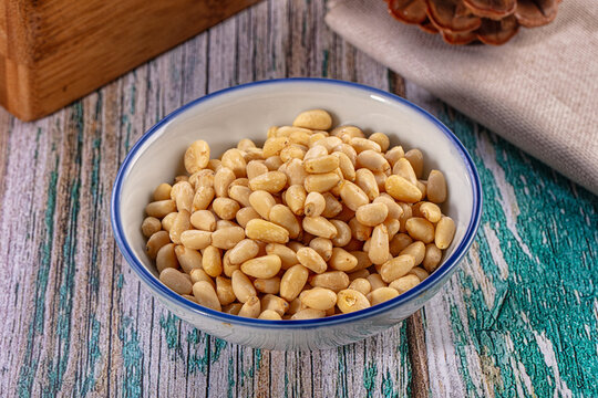 Close up of a ceramic bowl filled with shelled pine nuts on a rustic wooden surface with a pine cone