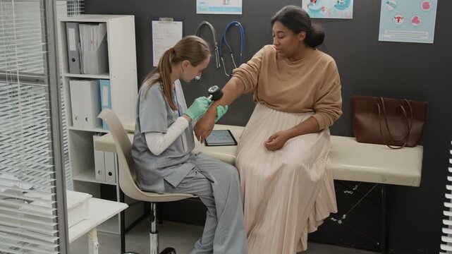 Doctor wearing scrubs and rubber gloves using dermatoscope while screening skin of female patients arm during medical checkup in clinic