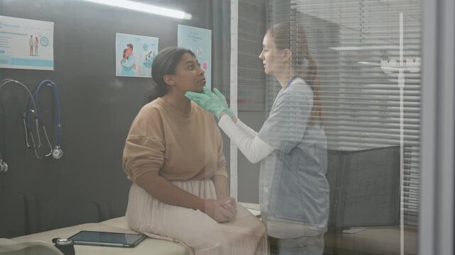 View through glass wall of endocrinologist palpating lymph nodes of womans neck during medical examination