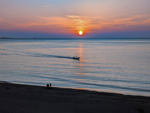 Beautiful landscape of a golden sunset ocean horizon. The vibrant orange sun warm across the sky , A small boat cruises in the middle ground, while two distant figures stand on the dark sandy beach
