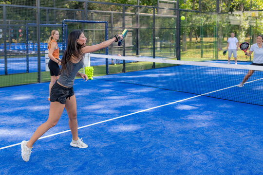 Woman hitting ball over net during padel doubles match, with opponents visible on the other side of the court.