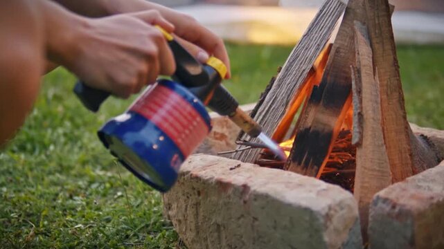 Closeup of woman hand using butane gas torch to ignite wooden logs in fire pit on grass, capturing fire starting process and outdoor camping activity in Savci Slovenia