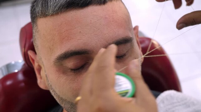 Barber uses thread to define and clean eyebrow lines. The scene highlights symmetry, accuracy, and refined grooming techniques for a polished look.